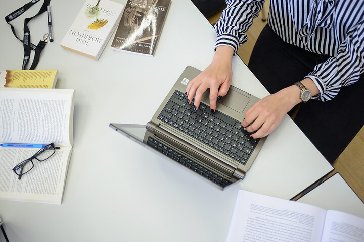 Laptop und aufgeklapptes Buch sind von oben zu sehen. Eine Person tippt auf dem Laptop.