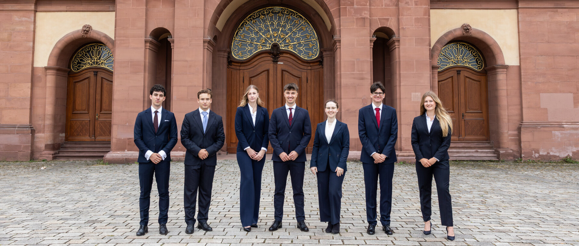 Eine Gruppe Studierender steht in Anzügen vor der Universität Mannheim.
