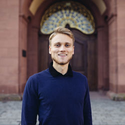 Tom Kranz trägt einen blauen Wollpullover und steht auf dem Ehrenhof der Uni Mannheim. Er hat kurze blonde Haare und einen Bart.