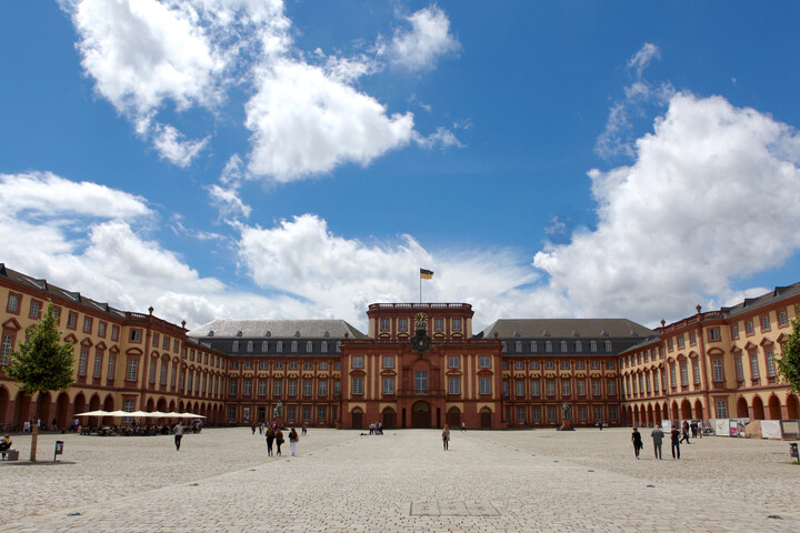 Das sandsteinfarbene Mannheimer Barockschloss mit Blick auf den Mittelbau bei strahlend blauem Himmel. Vereinzelt schlendern junge Menschen über das Kopfsteinpflaster auf dem Ehrenhof.