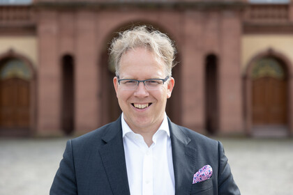 Portrait von Oliver Brand. Er trägt ein weißes Hemd sowie einen grauen Blazer und steht lächelnd auf dem Ehrenhof.