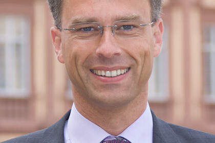 Portrait von Georg Bitter. Er trägt eine lila Krawatte sowuie einen grauen Blazer und steht lächelnd vor dem Schloss.