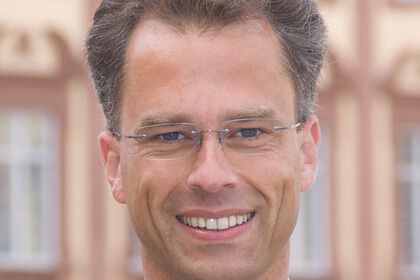 Portrait von Georg Bitter. Er trägt eine lila Krawatte sowuie einen grauen Blazer und steht lächelnd vor dem Schloss.
