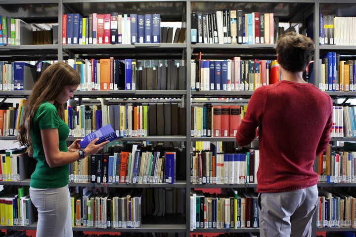 Zwei Studierende stehen vor einem Bücherregal einer Bibliothek 