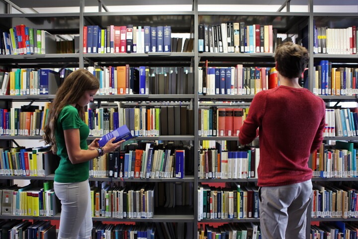 Zwei Studierende stehen vor einem Bücherregal einer Bibliothek 