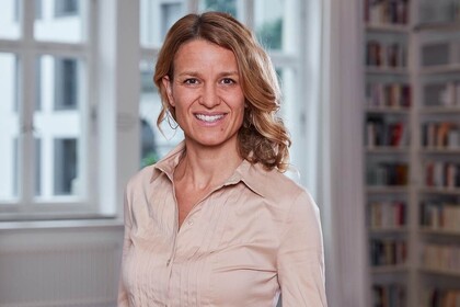 Portrait von Nadine Klass. Sie trägt eine beige farbene Bluse und steht lächelnd vor einem Fenster.