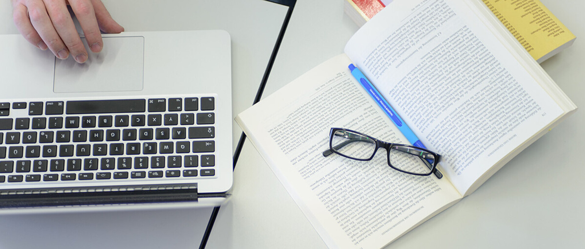 Aufgeklapptes Buch mit Brille und Stift, daneben Laptop mit Hand über Mousepad