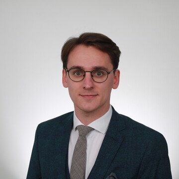 Wissenschaftlicher Mitarbeiter Laurin Krumwide. Er trägt ein weißes Hemd, eine gute Krawatte sowie eine dunkle Anzugsjacke.