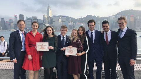 Eine Gruppe Studierender in Anzügen oder Kleidern stehen vor einem Fluss. Zwei Studierende halten ein Dokument in den Händen.