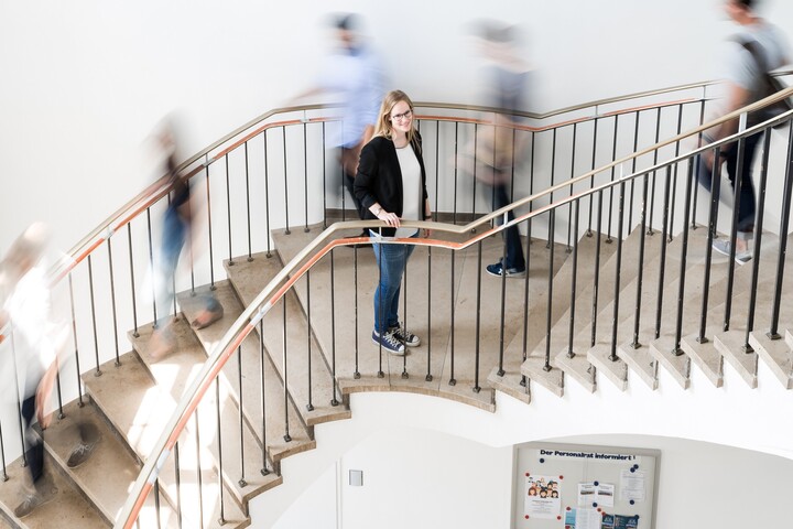 Dekoratives Bild, auf welchem eine Treppe zu sehen ist, auf welcher eine Person steht und deutlich zu erkennen ist. Andere Personen gehen und sind verschwommen.