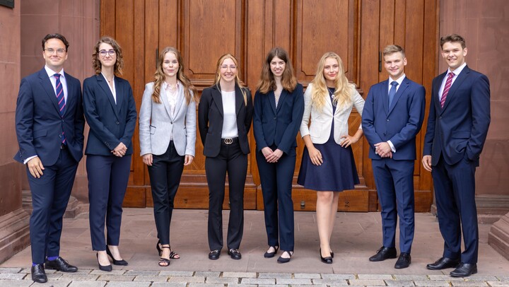 Eine Gruppe Studierender steht in Business-Kleidung lächelnd vor der Universität Mannheim.