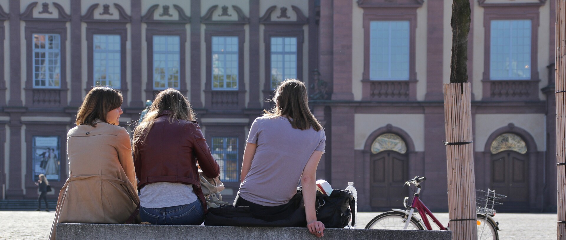 Drei Studierende sitzen mit dem Rücken zur Kamera auf einer Bank im Ehrenhof