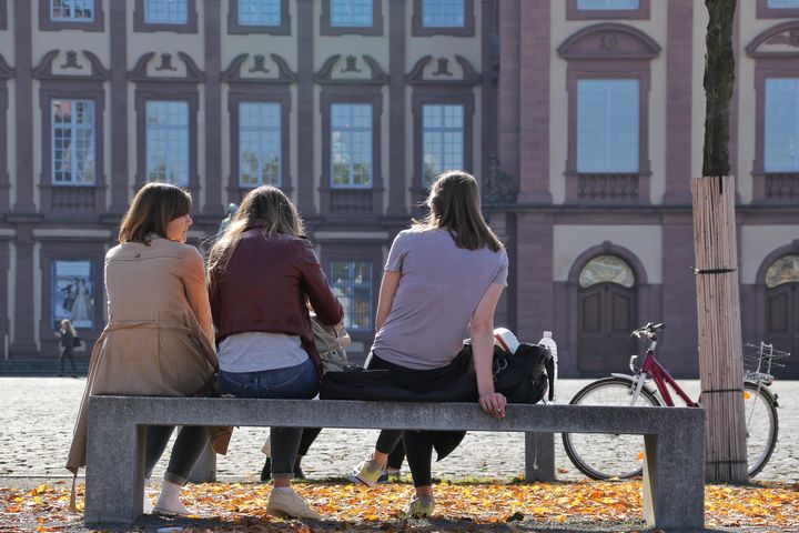 Drei Studierende sitzen auf einer Bank im Ehrenhof mit dem Rücken zur Kamera