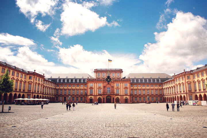 Das Barock-Schloss und der Ehrenhof der Universität Mannheim unter strahlend blauem Himmel. Das Schloss ist von unzähligen Fenstern, rotem Sandstein und einer gelben Fassade geprägt.